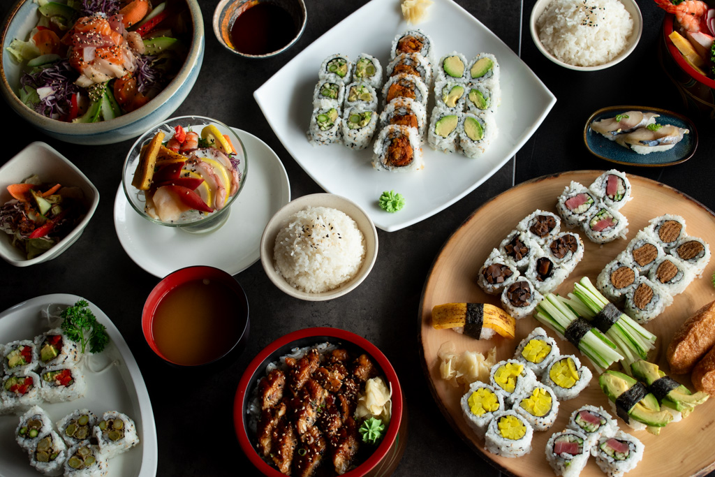 Table full of food from an asian restaurant called Three Kitchens. It includes a variety of sushi, hot plates, soups, rice, and salad.