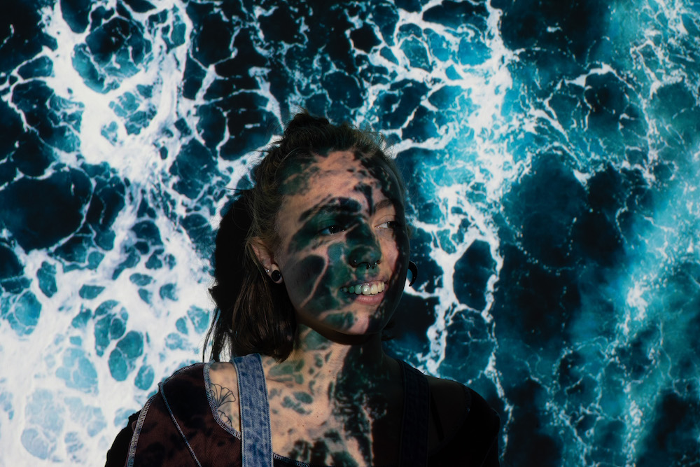 Girl standing looking towards the right side and smiles with a blue ocean projected on her face.